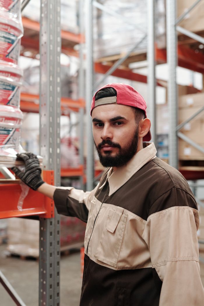 Services Portrait of a bearded man in a warehouse wearing a red cap and work uniform.
